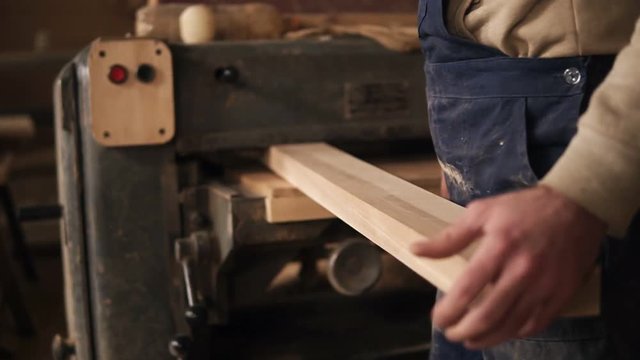A man works in a carpentry shop. He goes to the grinding machine and installs a long rail into it. Back view