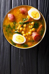 Stewed Potaje de garbanzos chickpeas, spinach, chorizo sausages, boiled eggs close-up in a bowl. Vertical top view