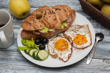 Croissant Sandwich with Fried Eggs, cucumbers and olives, Fruits and Fresh Vegetables and Cup of coffee on White Plate over Gray rustic Wooden Background.