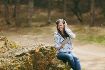 Naklejka na ściany i meble Young smiling woman in casual clothes sitting on stone listening music with headphones and tablet pc computer in city park or forest on green blurred background. Student lifestyle, leisure concept.