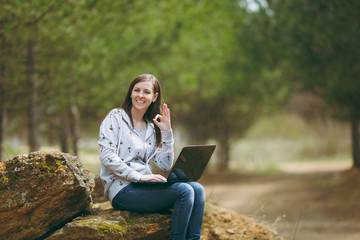 Obraz premium Young successful smart business woman or student in casual clothes sitting on stone using laptop and showing Ok sign in city park or forest working outdoors on green background. Mobile Office concept.