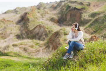 Young pensive relaxed beautiful woman in light casual clothes with headphones listening music sitting on grass with crossed legs looking aside on green field background. Lifestyle, leisure concept.