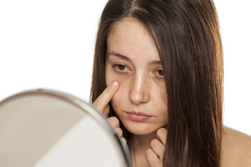 Unsatisfied young woman touch her face in mirror on white background