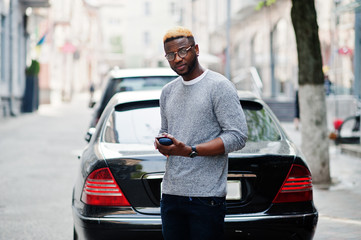 Fototapeta premium Stylish african american boy on gray sweater and glasses posed at street against black business car and speaking on phone. Fashionable black guy.