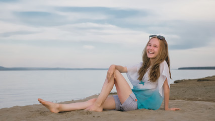 Fototapeta na wymiar One beautiful teenage girl with brown hair outside on a beautiful summer day. The woman is sitting on the beach. The sun was gone in the sunset. The girl is genuinely genuinely genuinely smiling and