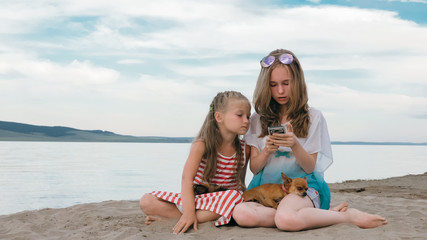 Fototapeta premium Two teenage are sitting on a sandy beach. They wear sunglasses. Sisters talk to each other and selfie on phone. They have two dogs. Girls correspond, playing, on the Internet.