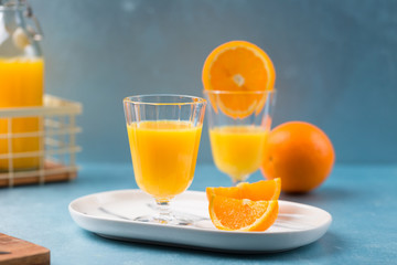 Glass of fresh orange juice with orange slices on white plate, blue background, cold summer refreshing nutritional beverage, blue orange color contrast, vitamin C healthy drink, copy space