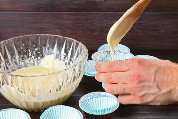 Hands pouring dough into molds for muffins on a wooden table.
