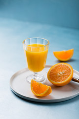 Glass of fresh orange juice with orange slices on white plate, blue background, cold summer refreshing nutritional beverage, blue orange color contrast, vitamin C healthy drink, copy space