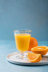 Glass of fresh orange juice with orange slices on white plate, blue background, cold summer refreshing nutritional beverage, blue orange color contrast, vitamin C healthy drink, copy space