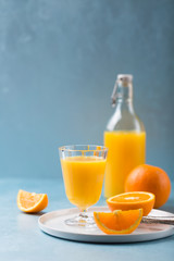 Glass of fresh orange juice with orange slices on white plate, blue background, cold summer refreshing nutritional beverage, blue orange color contrast, vitamin C healthy drink, copy space