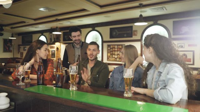 Dark-haired bearded young man is telling a story and gesturing emotionally while meeting with his friends in pub at counter. His mates are listening to him carefully.