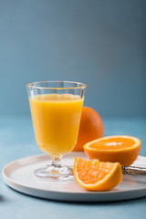 Glass of fresh orange juice with orange slices on white plate, blue background, cold summer refreshing nutritional beverage, blue orange color contrast, vitamin C healthy drink, copy space