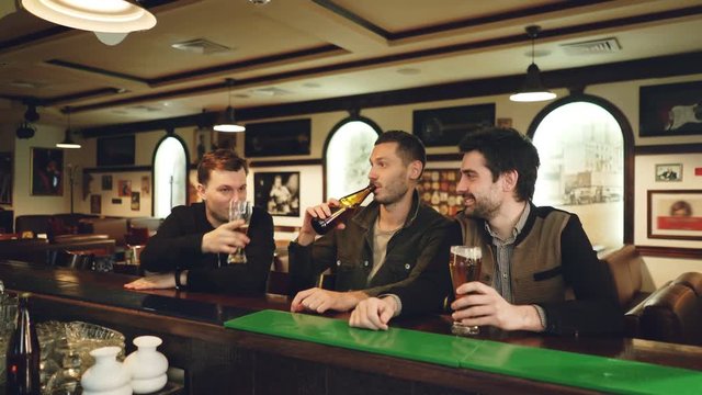 Three young men in casual clothes are drinking beer in fancy sports bar, clanging glasses and bottles and talking while sitting at counter. Guys having night out concept.