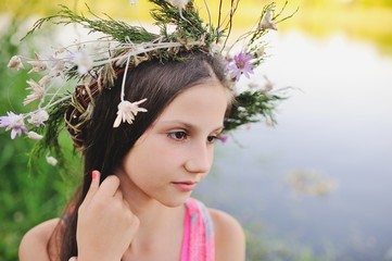 Child girl in a wreath of wild flowers on a background of a river and fresh green grass. Midsummer day. Celebration of the holiday of Ivan Kupala