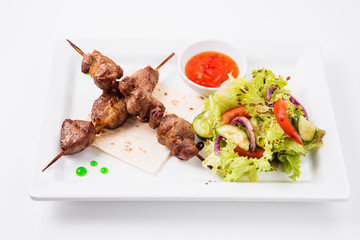 Shish kebab on bamboo skewers lies on a pita. Near sauce and fresh vegetable salad. White background (top view)