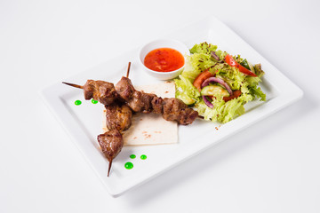 Shish kebab on bamboo skewers lies on a pita. Near sauce and fresh vegetable salad. White background