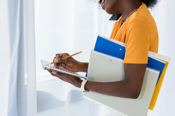 cropped shot of african american woman with folders talking on smartphone and making notes in notebook at home
