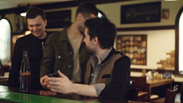 Young handome man is using smartphone at bar counter when his male friends are coming, watching screen and discussing something. Technology communication concept