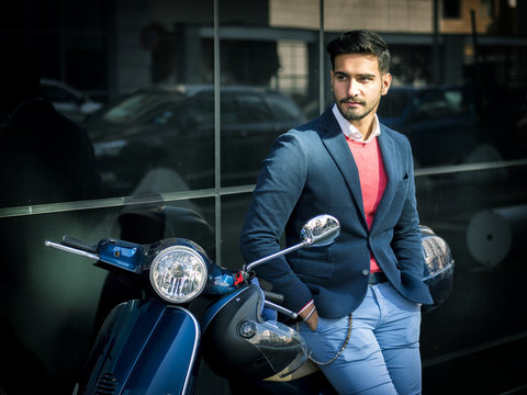Young stylish man in suit and sunglasses standing by his classic Italian scooter on empty road in sunny city.