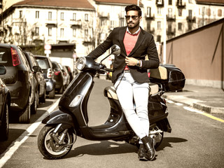Fototapeta na wymiar Young stylish man in suit and sunglasses standing by his classic Italian scooter on empty road in sunny city.