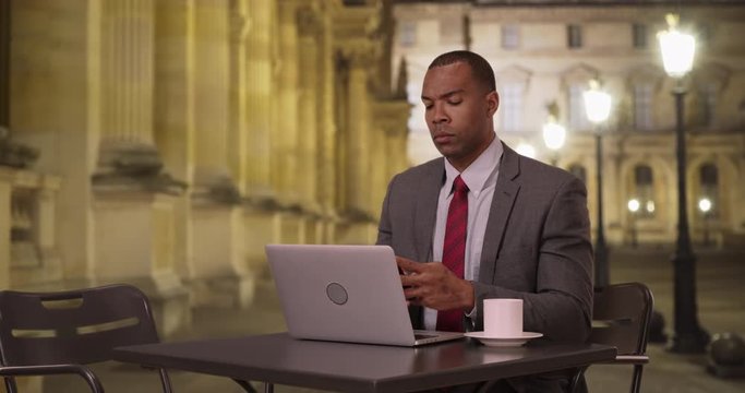 African-American businessman sitting at a public table using his laptop and smartphone for work, Determined young professional working hard on his computer on a night in Paris, 4k