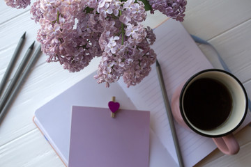 Notepad, pencils, lilac in vase and a cup of coffee on a white wooden desk. Inspirational workplace.