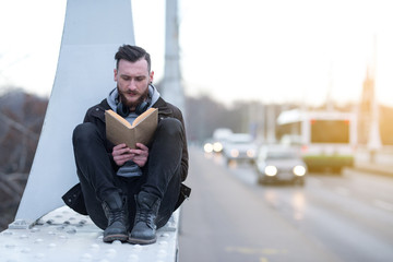 A handsome young hipster man sitting on a bridge and reading a book.
