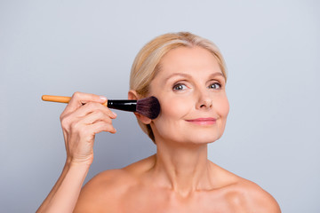 Portrait of charming woman with smooth soft perfect skin applying rouge with brush on cheek looking at camera isolated on grey background