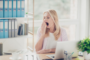 Pretty, attractive, stylish, modern, aged woman want to sleep, holding hand near open mouth, yawning with close eyes, sitting at desk in workplace