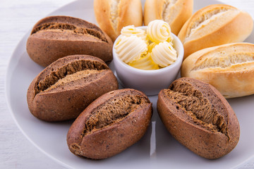 Roll of black bread, is served with butter on a white plate and a white wooden background