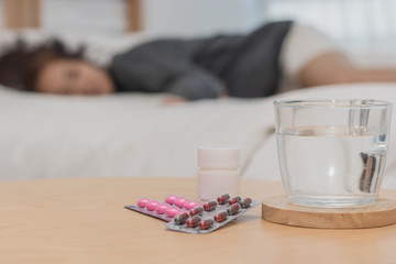 Closeup on pills and glass of water on table and Asian women sleep sick on bed in bed room
