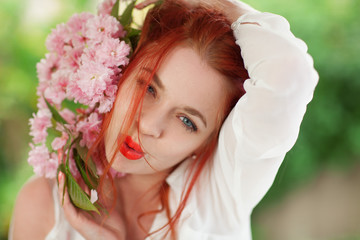 Naklejka premium Beautiful young woman with red hair having fun standing in the garden with cherry blossom branch and perfect make up