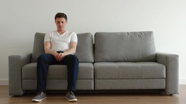 Sad young adult in a white t-shirt and a jeans sits on a sofa