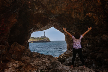 Woman expressing joy in cave beside the sea. Lighthouse in the background