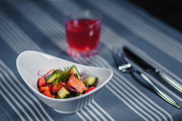 Table in the restaurant. On the table is a salad in a white plate, a knife, a fork, a glass with red juice. On the table is a blue tablecloth in strips.