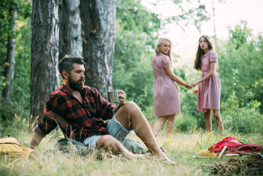 Friends camping in forest. Two girls holding hands look back at man sitting on grass. Side view bearded guy sitting on green meadow