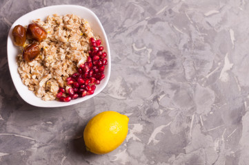 Cooked yummy warm oatmeal and heap of ripe fresh pomegranate seeds and three dried dates in white ceramic bowl near whole lemon on worn gray scratched concrete. Top view with copy space