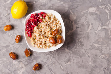 Fototapeta na wymiar Cooked yummy warm oatmeal and heap of ripe fresh pomegranate seeds and three dried dates in white ceramic bowl near whole lemon and scattered dates on worn gray scratched concrete with copy space