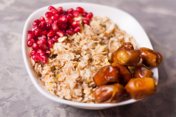 Cooked yummy warm oatmeal and heap of ripe fresh pomegranate seeds and lot of dried dates in white ceramic bowl on worn gray scratched concrete