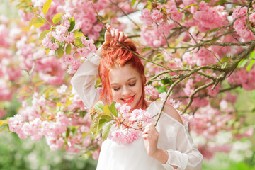 Beautiful young woman with red hair having fun standing in cherry blossom tree, springtime garden...