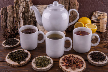 A cup with green tea, a cup with red tea and a cup with black tea, a big teapot, lemons on a dark wooden background
