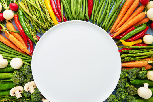 Vegetables background with empty white plate in the middle. Carrots, tomatoes, asparagus, broccoli, chilli pepper, green beans. Top view, copy space. Organic food.