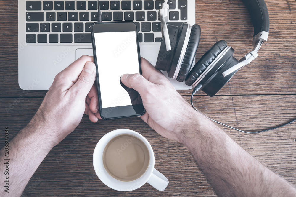 Wall mural man using smartphone on old wooden table with laptop computer, headphones and cup of coffee