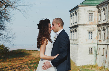 Wedding couple is kissing outdoors in sunny autumn day. Beautiful nature and renaissance palace on background. Bride is satin lace dress is holding groom hand. Old castle architecture.
