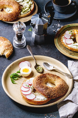 Variety of homemade bagels with sesame seeds, cream cheese, pesto sauce, eggs, radish, herbs served on ceramic plate with ingredients and coffee above over blue texture background. Top view, space.