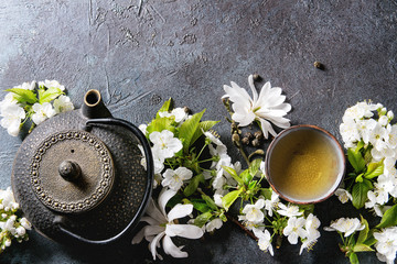 Traditional ceramic cup of hot green tea with black iron teapot, spring flowers white magnolia and cherry blooming branches over dark blue texture background. Top view, copy space.