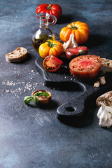 Variety of red and yellow organic tomatoes with olive oil, garlic, salt and bread for salad or bruschetta on wooden cutting board over blue texture background. Copy space.