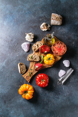 Variety of red and yellow organic tomatoes with olive oil, garlic, salt and bread for salad or bruschetta on wooden cutting board over blue texture background. Top view, space.