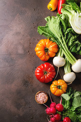 Variety of wet raw fresh organic colorful vegetables tomatoes, radish with leaves, fennel, paprika with bowl of pink salt for salad over dark brown texture background. Top view, space.
